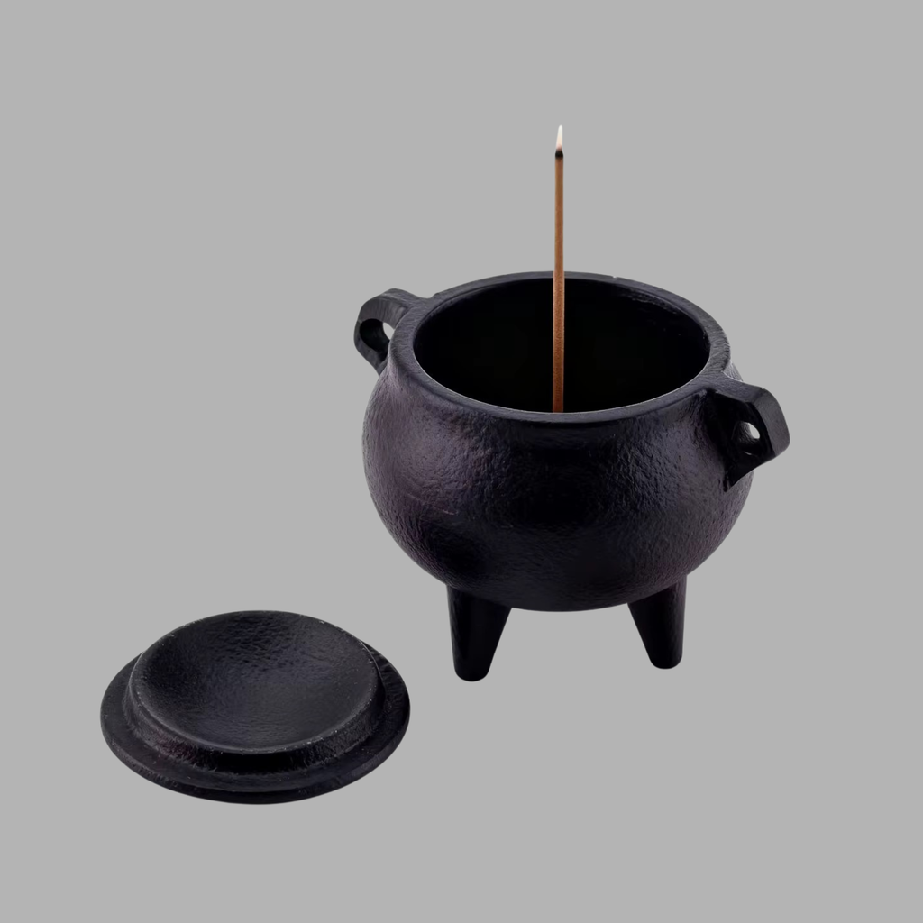 A black cast-iron incense burner with a lid placed beside it, shown on a plain gray background. A single incense stick stands upright inside the pot.
