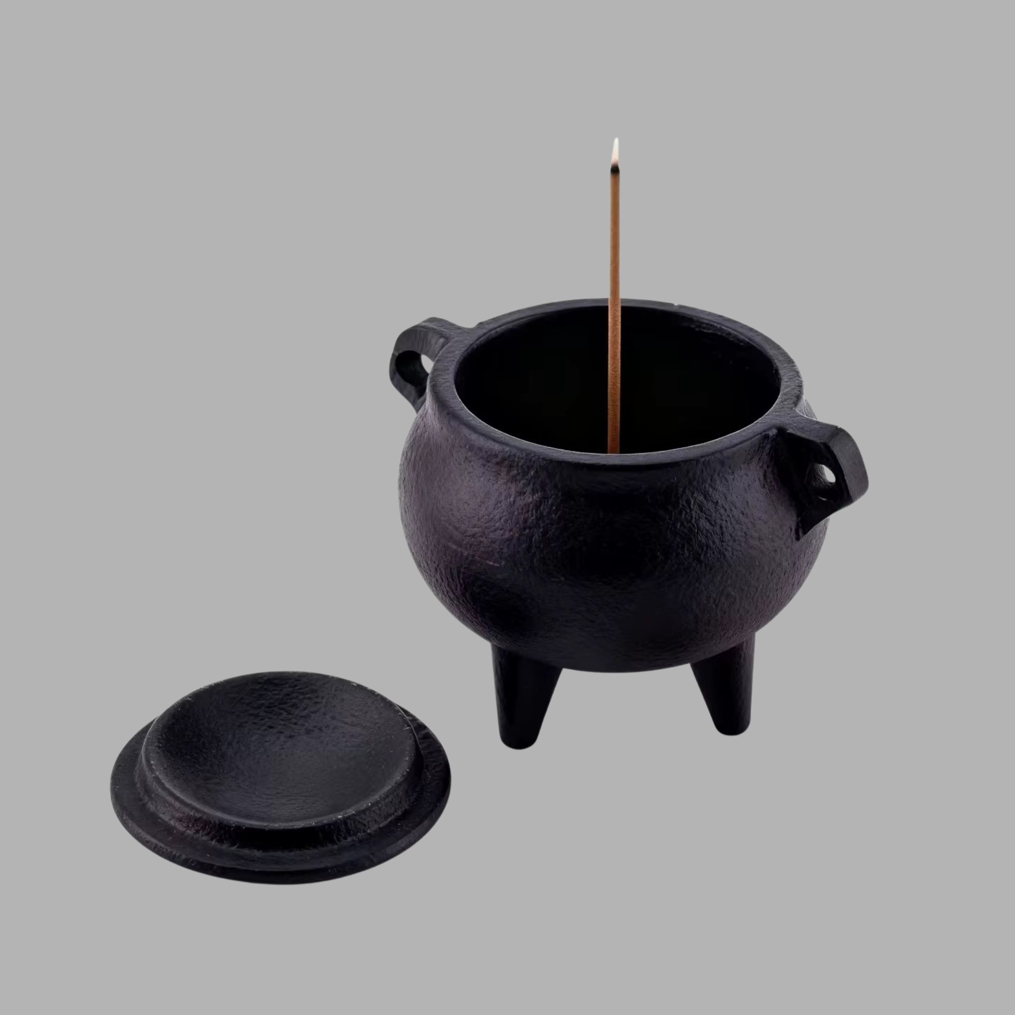 A black cast-iron incense burner with a lid placed beside it, shown on a plain gray background. A single incense stick stands upright inside the pot.